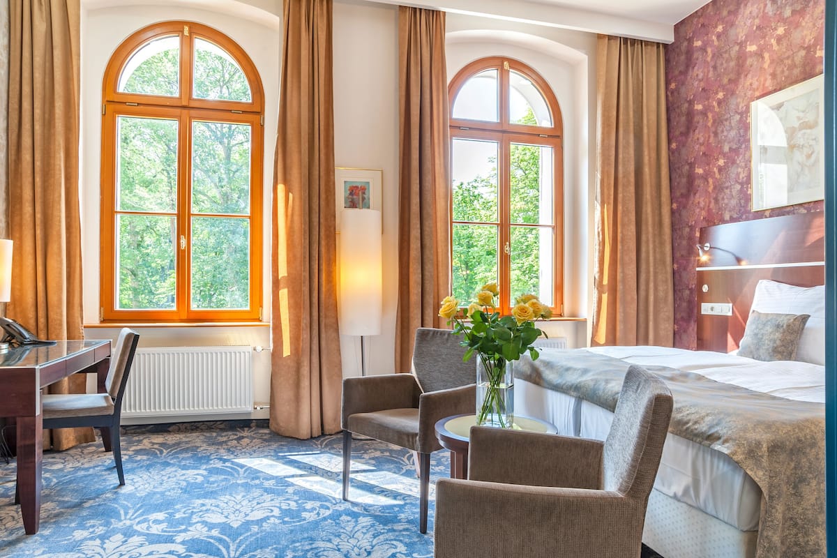 Ein Hotelzimmer mit einem großen Bogenfenster, einem Schreibtisch mit Stuhl, einem Bett mit Kopfteil und einer Vase mit Blumen.