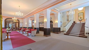Un hall d’hôtel avec un tapis rouge, des espaces aménagés avec des sièges, des lustres et un escalier.