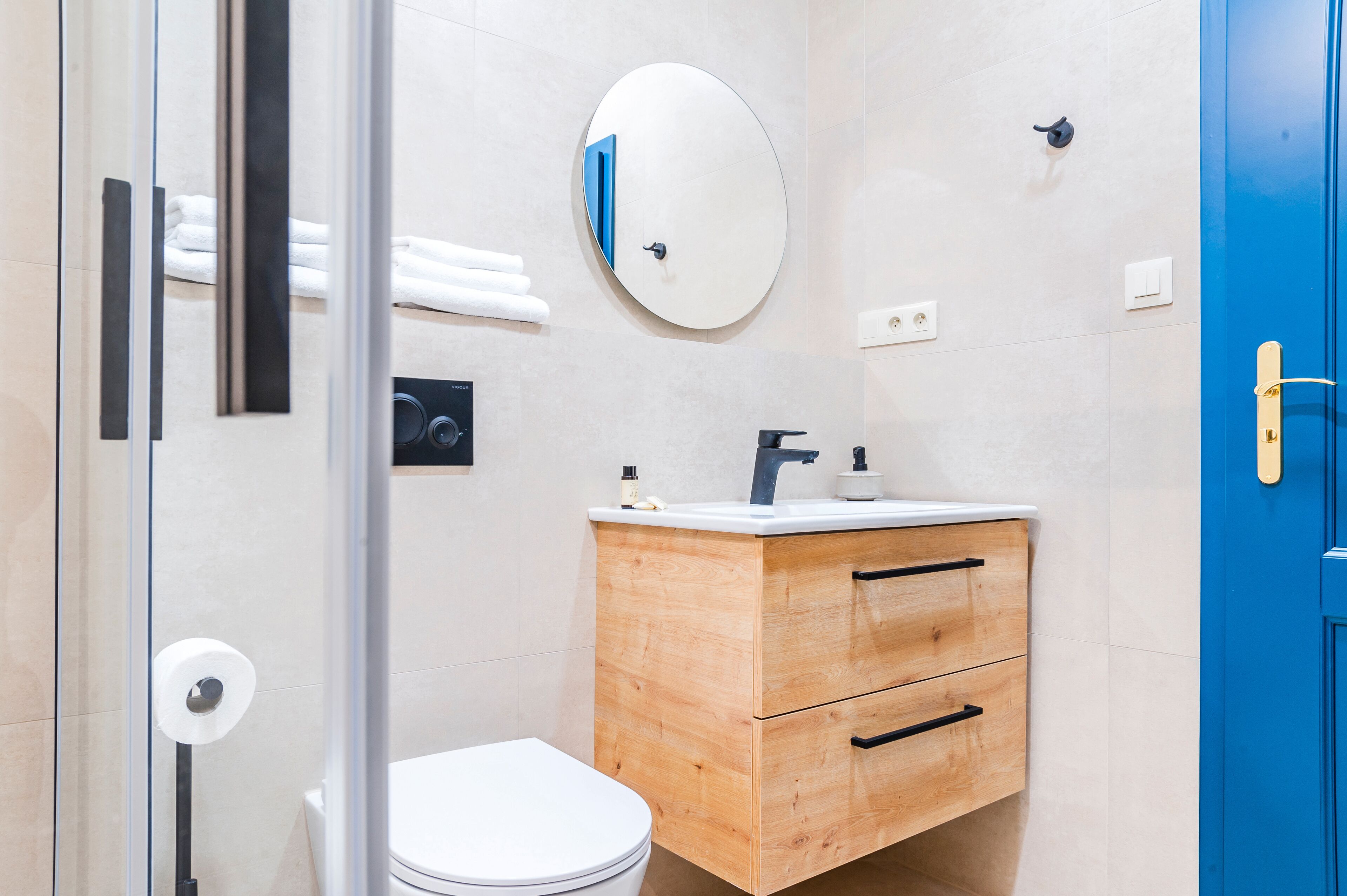 A bathroom with a white toilet, wooden vanity with two drawers, a round mirror, and a blue door.