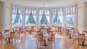 Un espace repas avec des tables et des chaises rondes, de grandes fenêtres avec des rideaux et un lustre.