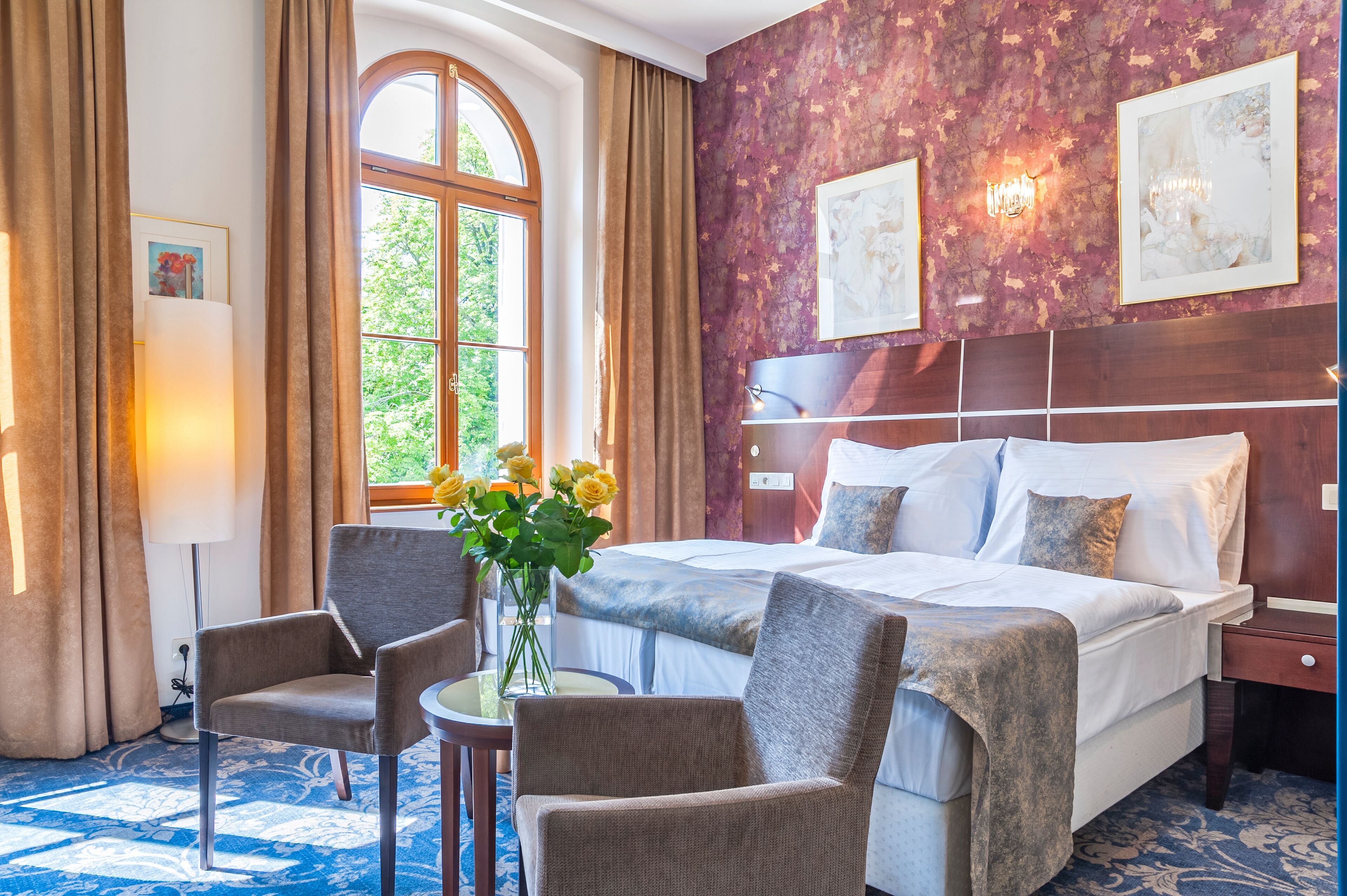 A hotel room with a large bed, two armchairs, a small table with a vase of flowers, and a large window with curtains.