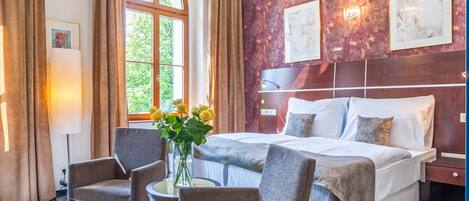 A hotel room with a large bed, two armchairs, a small table with a vase of flowers, and a large window with curtains.