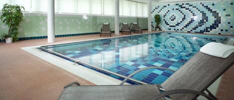 An indoor swimming pool with tiled walls, lounge chairs, and potted plants.