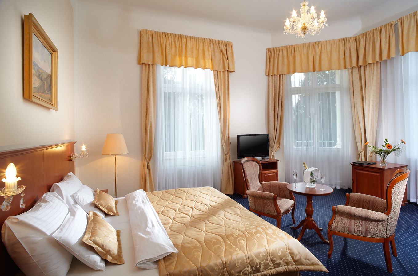 A hotel room with a large bed, two chairs, a small table, a TV, and a chandelier.