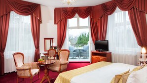 A hotel room with a large bed, two armchairs, a small table with a fruit bowl and wine glasses, a television on a wooden cabinet, and a balcony with outdoor seating. - Imperial Spa Hotel (Frantiskovy Lazne)