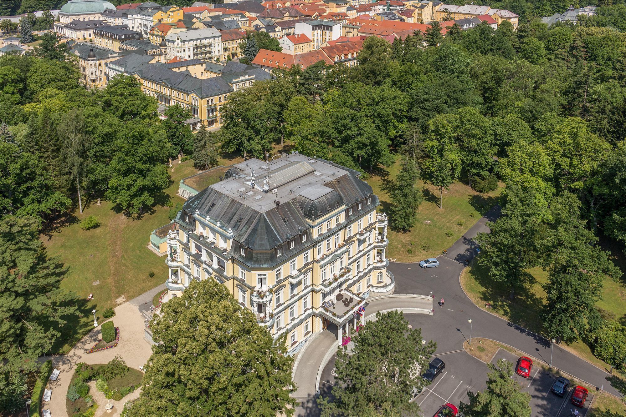 An aerial view of a large, historic building with a prominent dome, surrounded by lush greenery and a parking area with several cars.