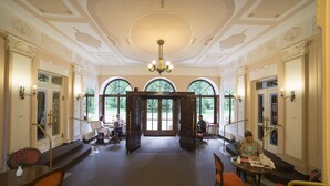 Un lobby espacioso con una escalera central, techo ornamentado y amplias ventanas en arco que ofrecen vistas al jardín.