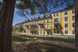 A row of yellow multi-story buildings with balconies, a paved walkway, and a tree in the foreground. - Dr. Adler Spa Hotel (Frantiskovy Lazne)
