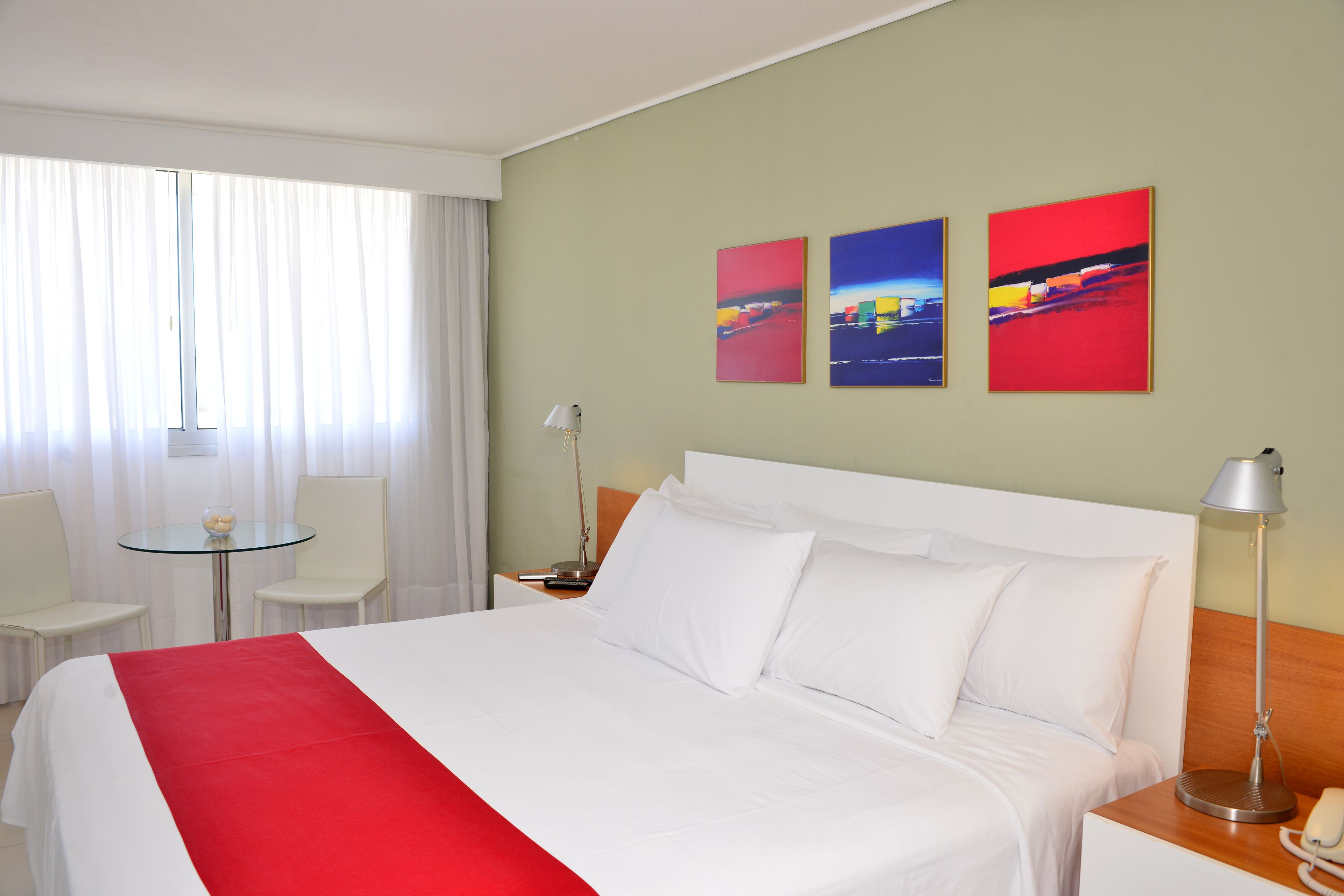 A hotel room with a large bed, white curtains, a small round table with chairs, and three colorful paintings on the wall.