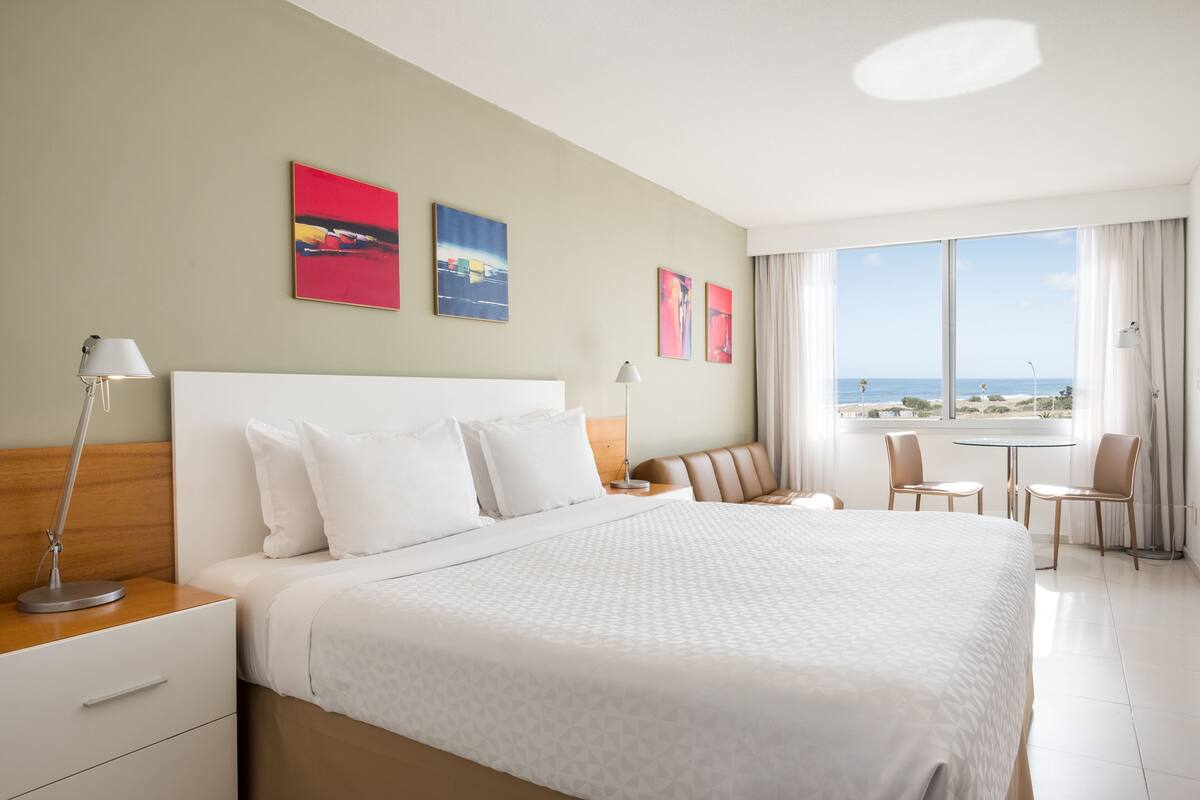 A hotel room with a large bed, a desk, a chair, a small table, and a view of the ocean.
