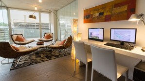 A modern office space with two monitors, wicker chairs, a white desk, and a large abstract painting on the wall. - UY Proa Sur Hotel (La Paloma)