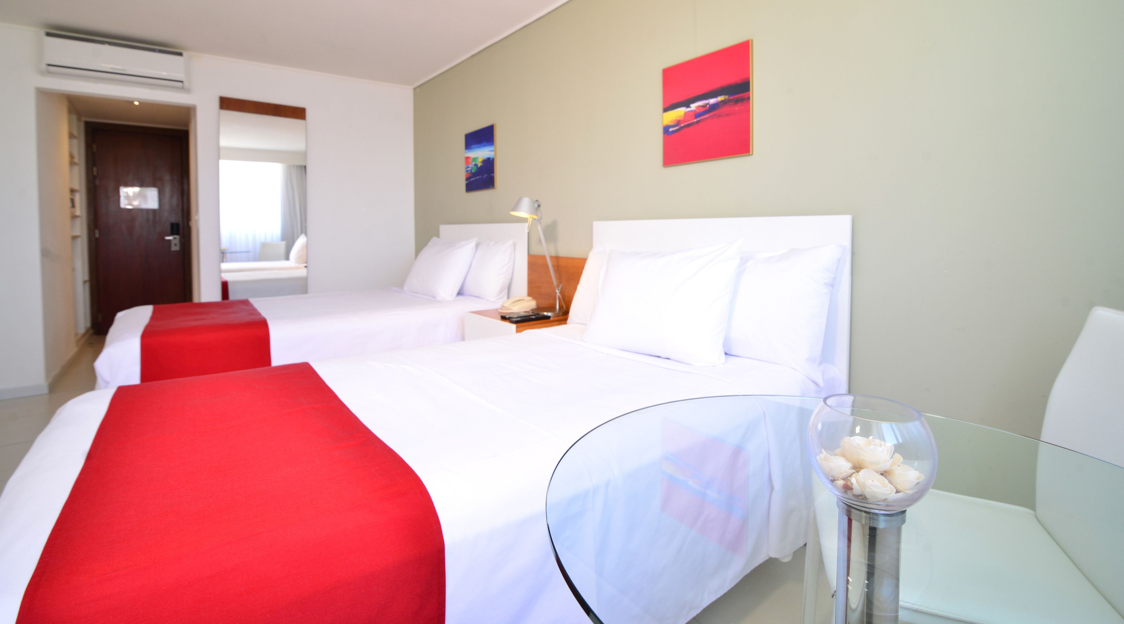 Habitación de hotel con dos camas, una mesita de vidrio con un florero, y una mesita de noche con una lámpara.