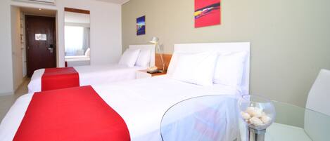 Habitación de hotel con dos camas, una mesita de vidrio con un florero, y una mesita de noche con una lámpara.