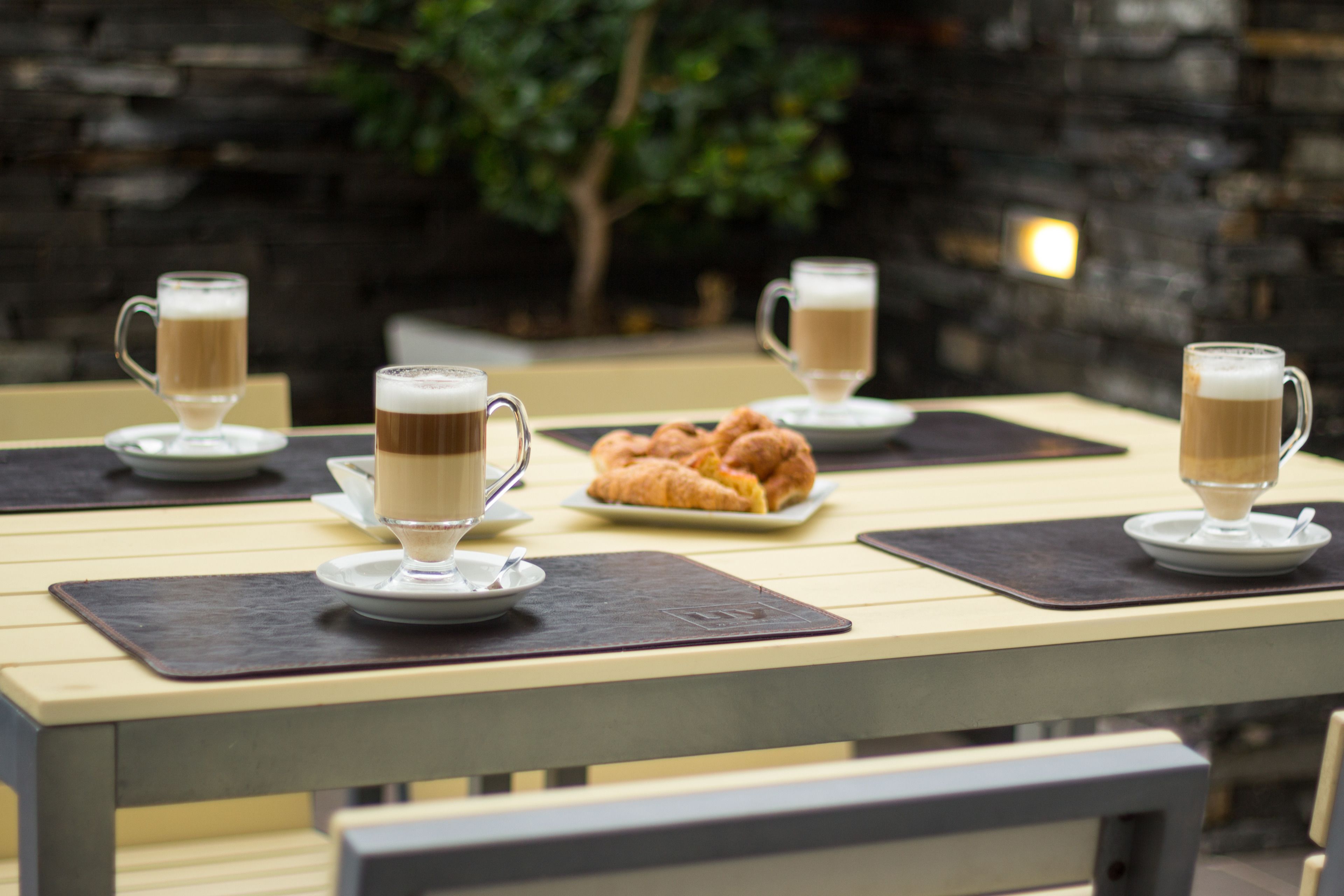 A table with four coffee cups and a plate of croissants, set against a stone wall background.