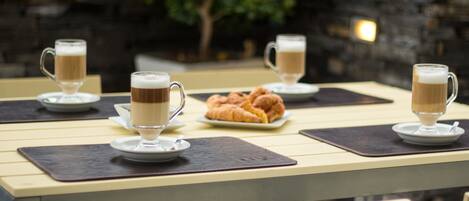 A table with four coffee cups and a plate of croissants, set against a stone wall background.