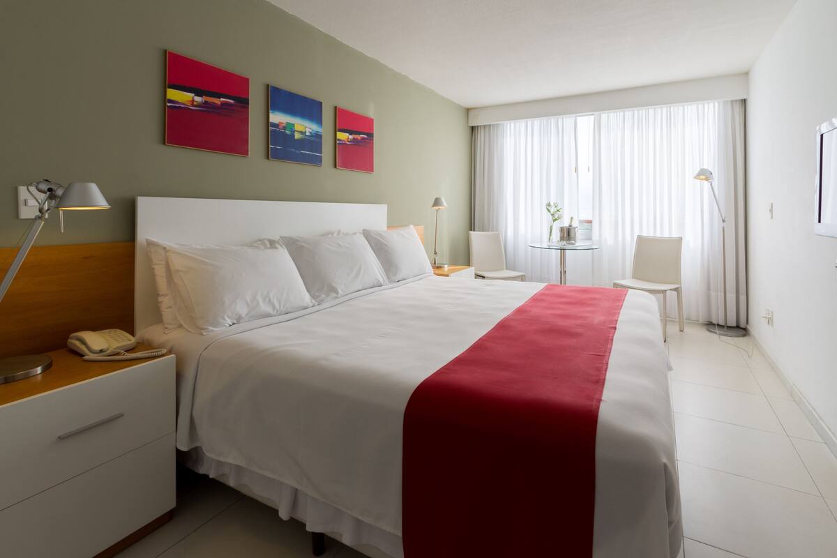 A hotel room with a large bed, bedside tables, a desk, and a chair. There are three paintings on the wall, a window with curtains, and a lamp on the desk.
