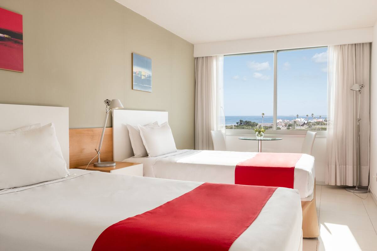 A hotel room with two beds, a view of the sea, a small table with a vase, and a window with curtains.