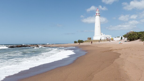 A beach with a white lighthouse, sandy shore, and ocean waves. - UY Proa Sur Hotel (La Paloma)