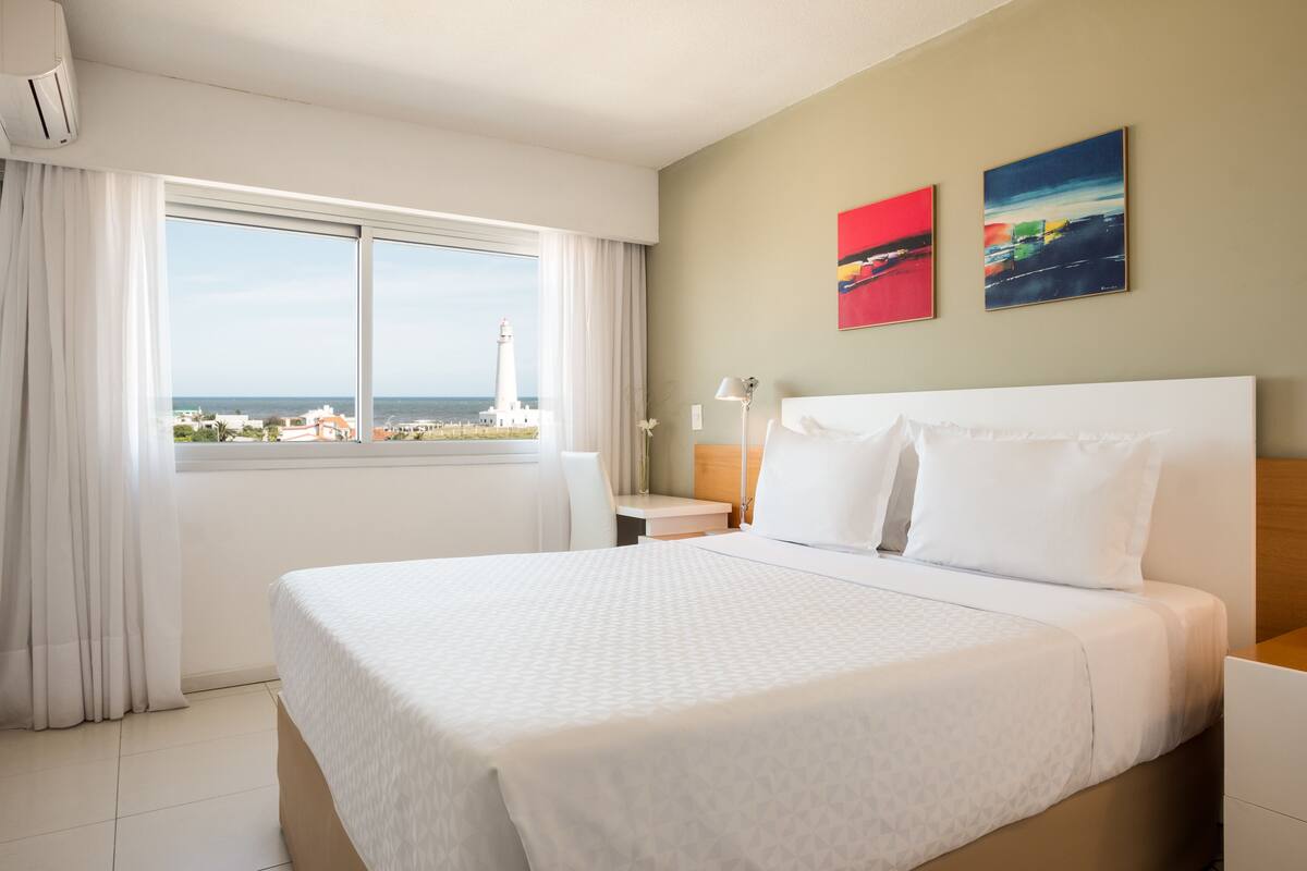 A hotel room with a large bed, a desk, a chair, and a view of the sea and a lighthouse.