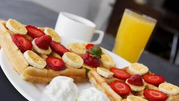 Un plato de desayuno con waffles cubiertos de fresas y rodajas de plátano, una cucharada de crema batida y un vaso de jugo de naranja.
