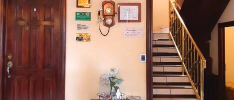 A reception area with a wooden door, a desk with a telephone, framed certificates, and a staircase.