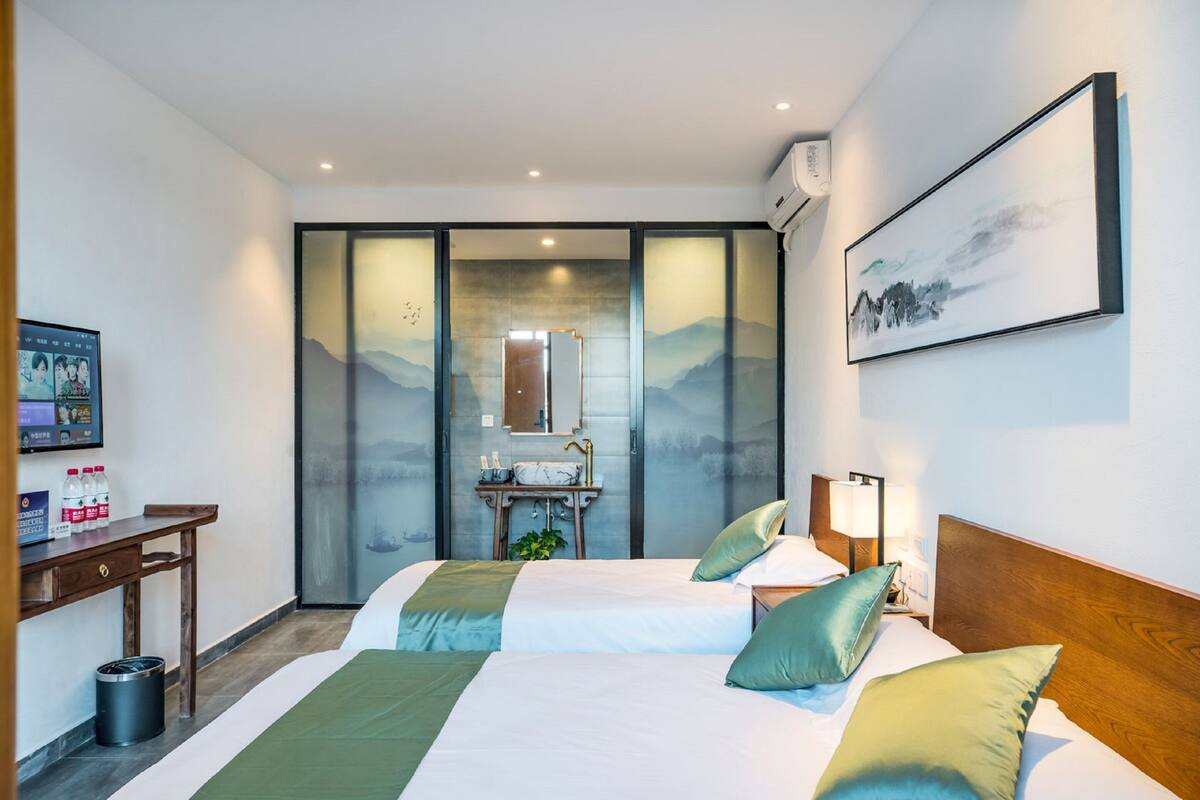 A hotel room with two beds, a desk, a mirror, a sink, and a painting on the wall.