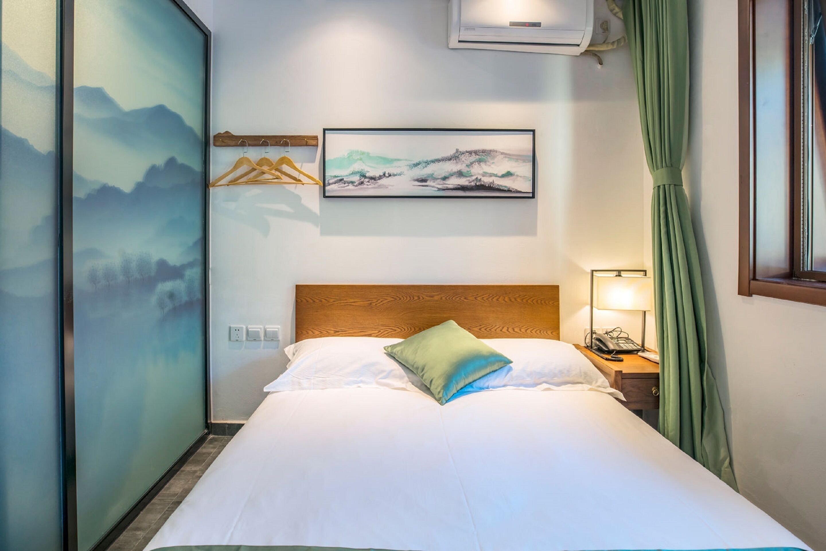 Een net opgemaakt bed met witte lakens en één groen kussen, een houten hoofdbord, een schilderij van een berglandschap aan de muur, een houten kleerhanger en een nachtkastje met een lamp en telefoon.
