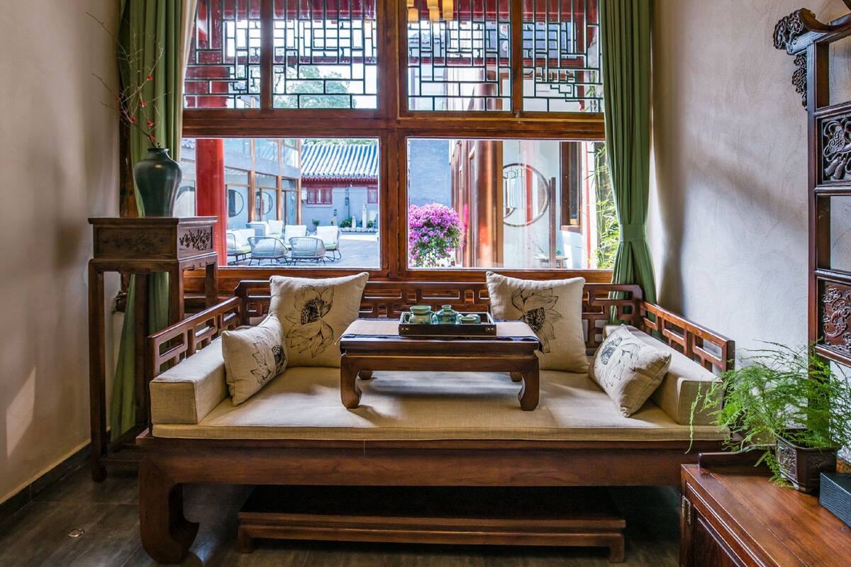 A traditional wooden sofa with cushions, a small wooden table, and a view of a traditional building through a large window with green curtains.