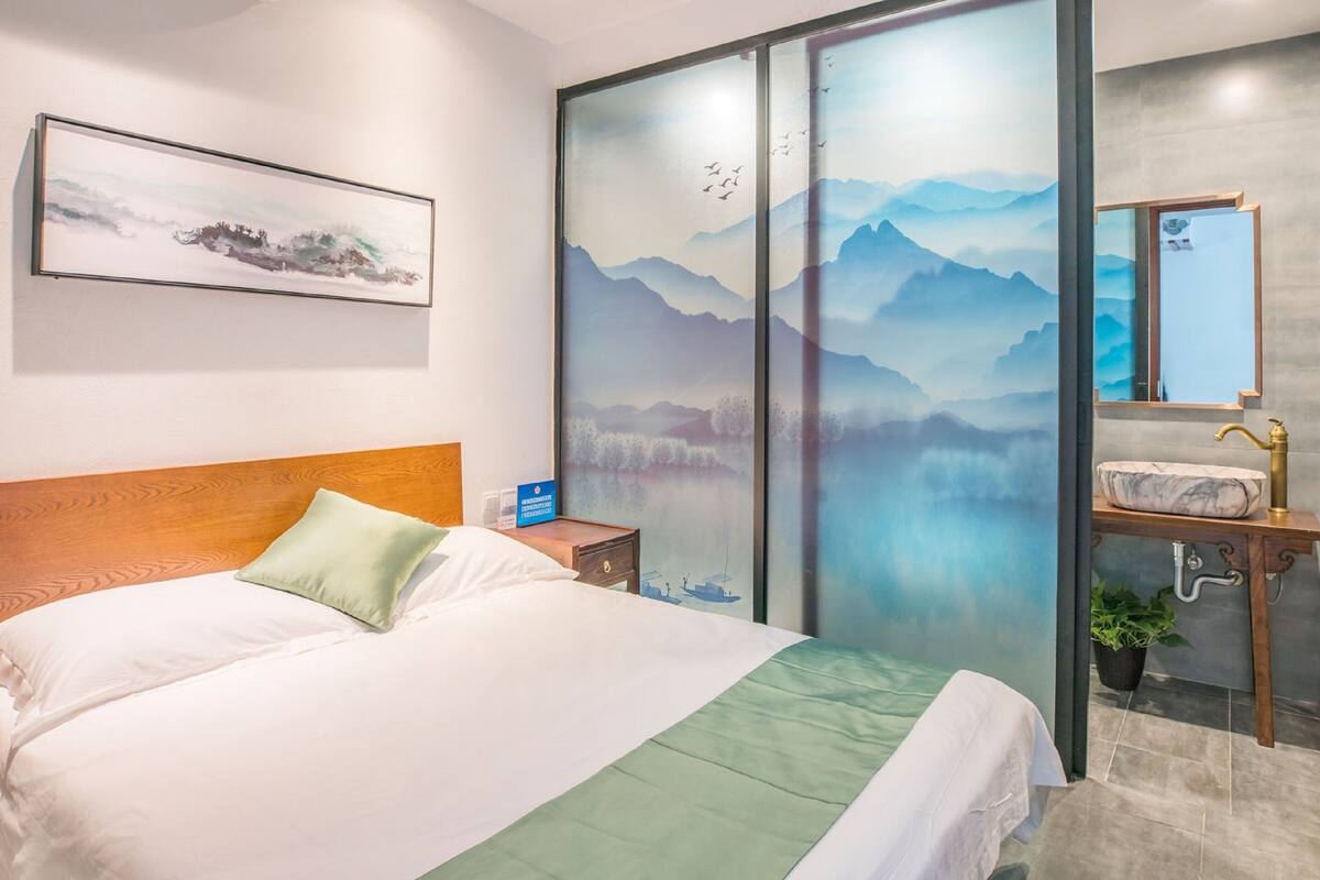 A hotel room with a bed, a nightstand, a wall-mounted painting, a bathroom with a sink and mirror, and a sliding glass door with a mountain view.