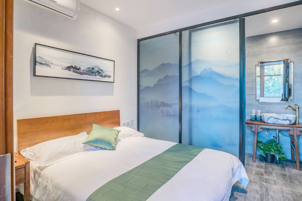 A modern hotel room with a large bed, a wooden headboard, a painting on the wall, a sliding glass door with a mountain view, and a small bathroom with a sink and mirror.