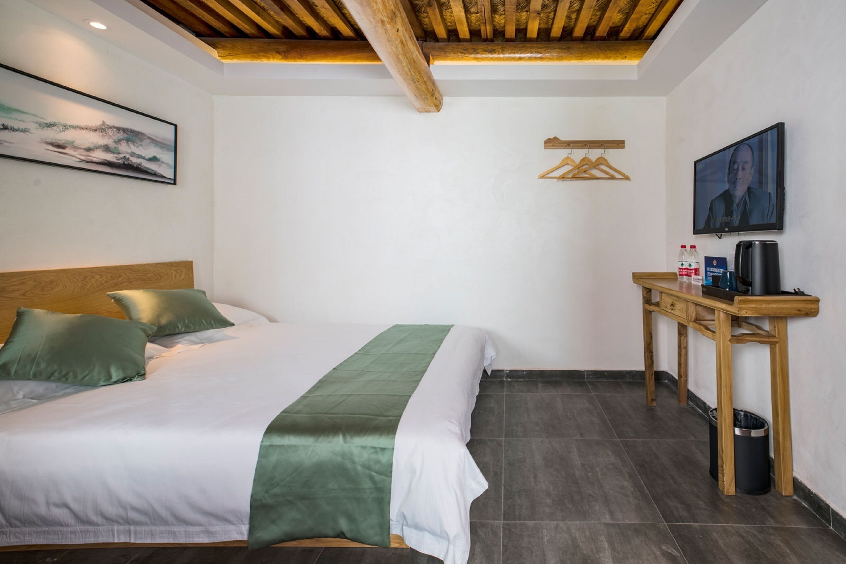 A hotel room with a bed, a wooden headboard, a green and white bedspread, a wooden nightstand with a coffee maker, a trash can, and a painting on the wall.