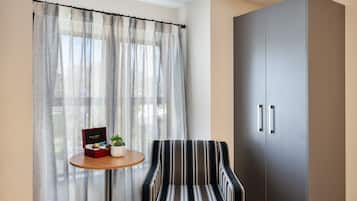 Ein Zimmer mit einem gestreiften Sessel, einem kleinen runden Tisch mit einer Schachtel und einer Pflanze, einem Kleiderschrank und durchsichtigen Vorhängen.