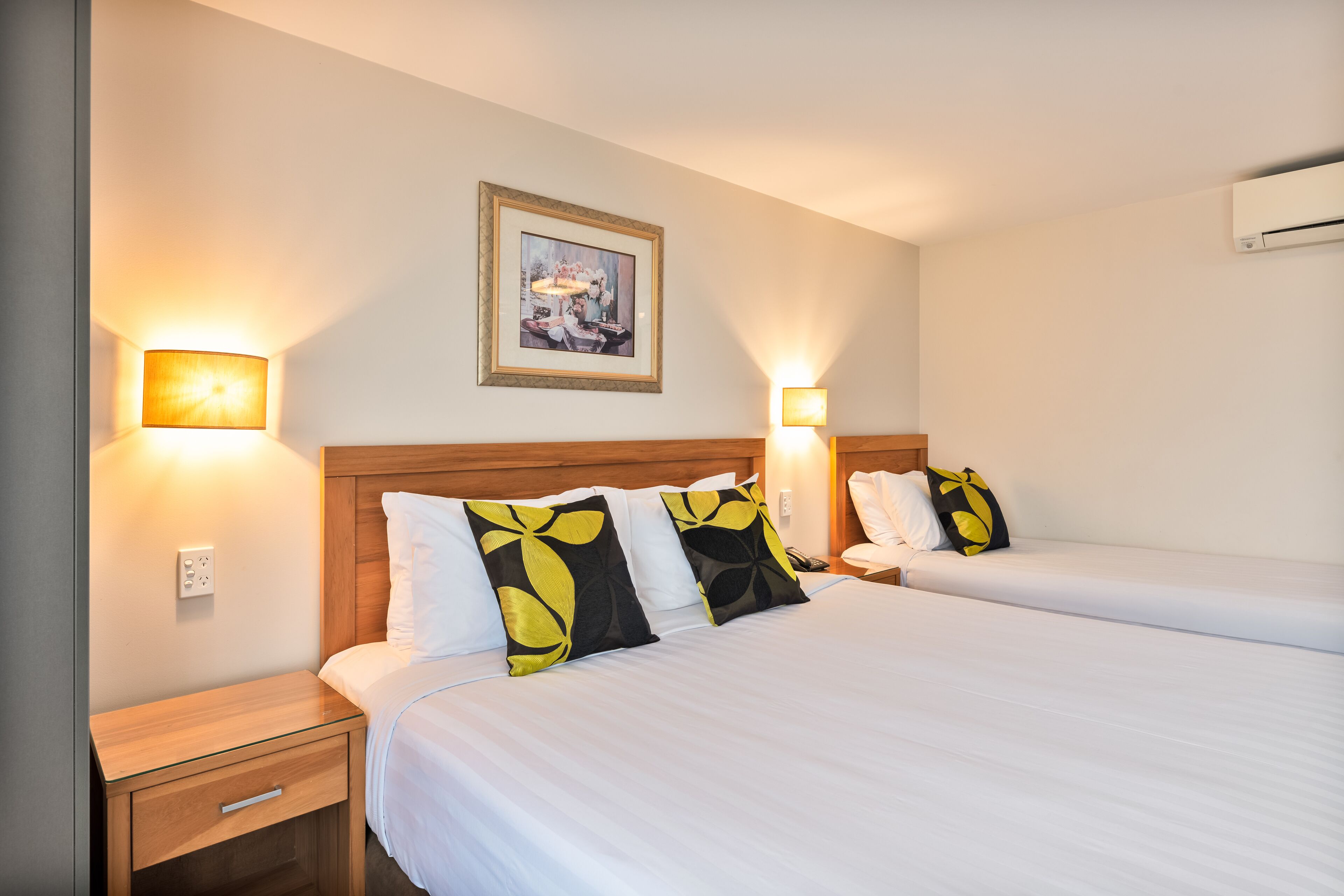 Une chambre d’hôtel avec deux lits, une tête de lit en bois, des lampes de chevet, un tableau encadré au mur et un appareil de climatisation.