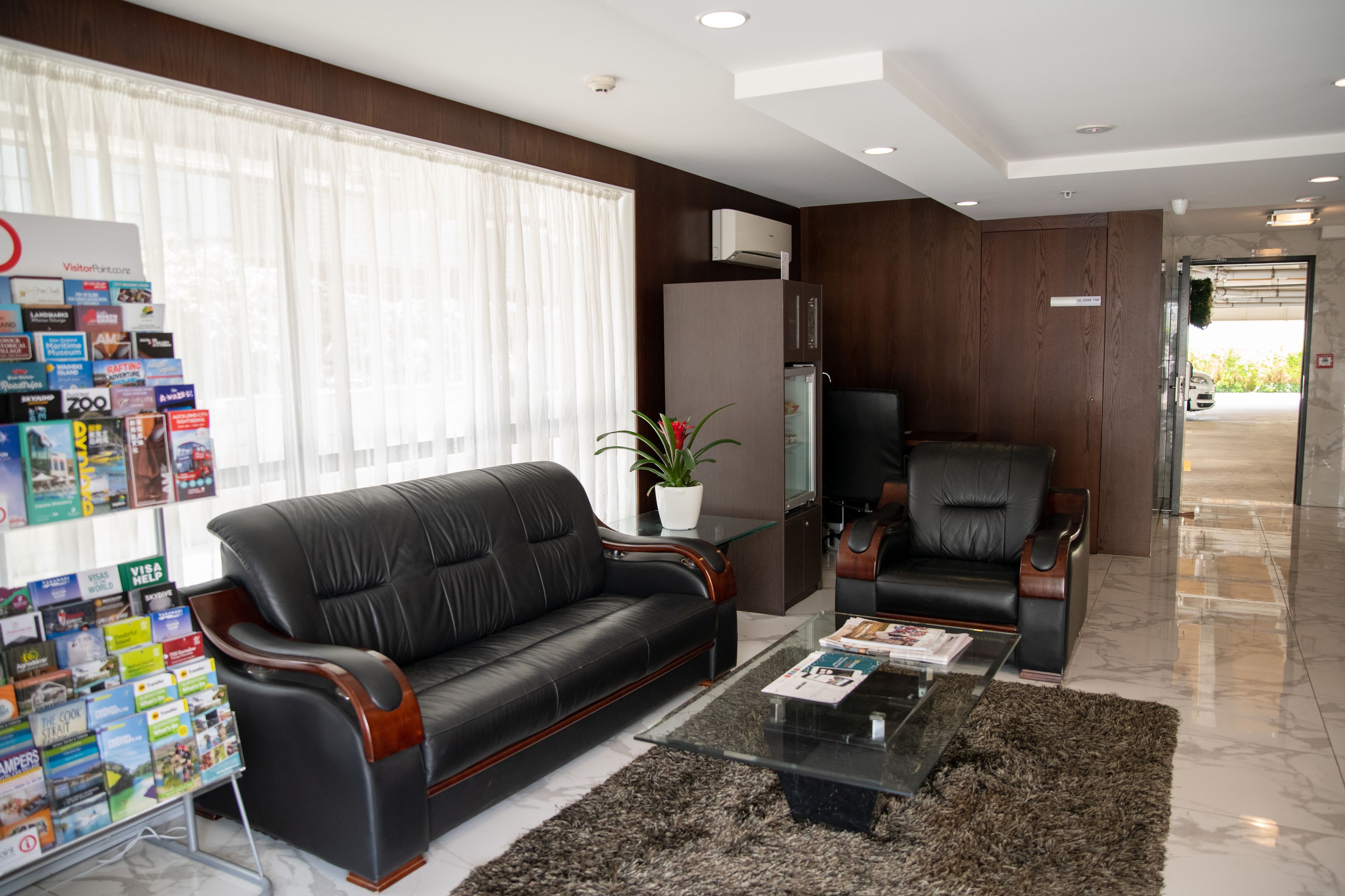 Una sala de estar moderna con un sofá de cuero negro, una mesa de centro de vidrio y una planta en maceta. Hay un mostrador de recepción con folletos y un cartel que indica los servicios.