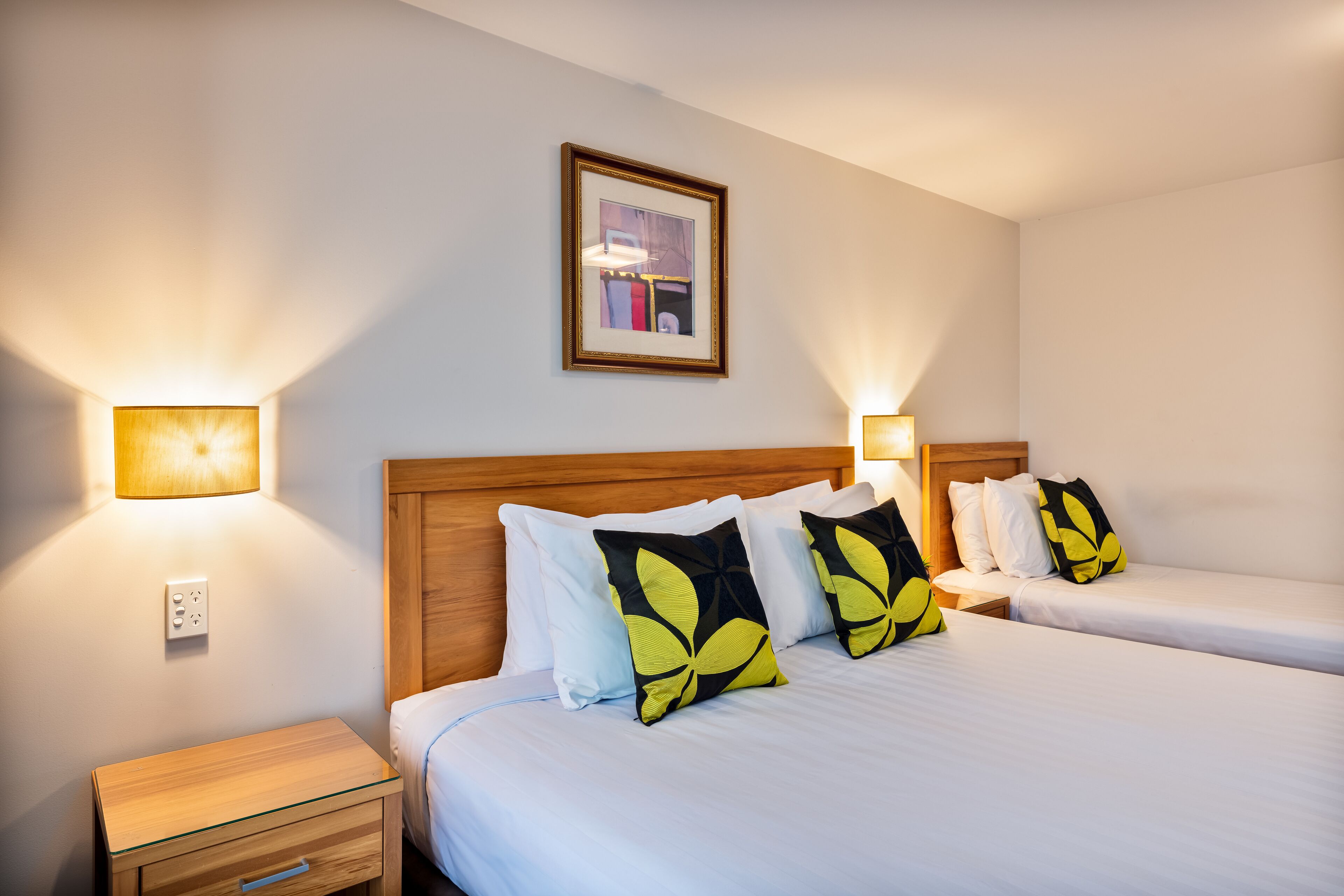 A hotel room with two beds, a wooden headboard, bedside lamps, a framed picture on the wall, and a small wooden nightstand with a drawer.
