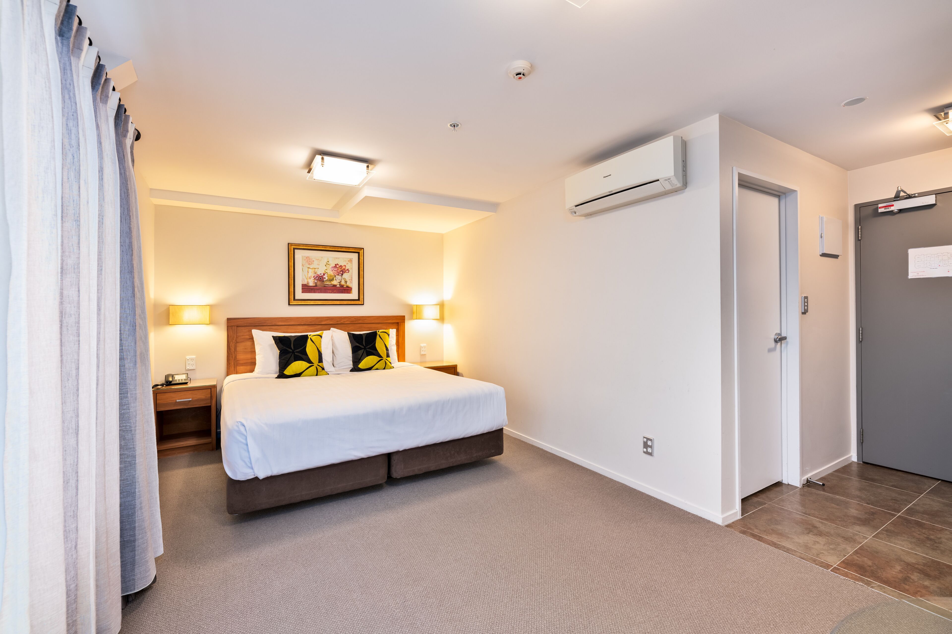 A hotel room with a bed, bedside lamps, an air conditioning unit, and a door.