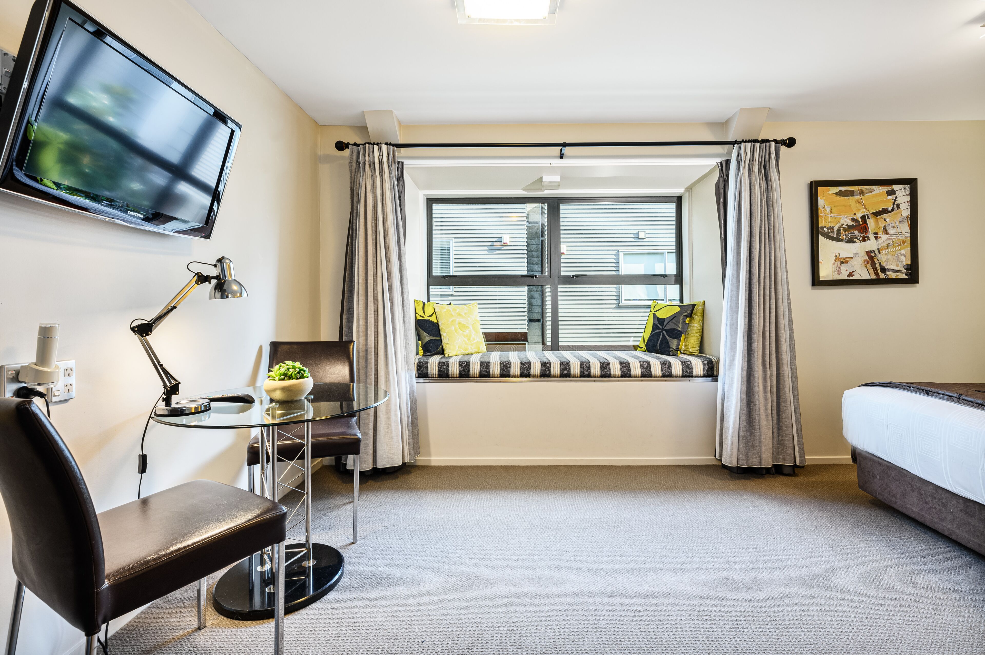 Et hotelværelse med seng, skrivebord med stol, fjernsyn ophængt på væggen, et vindue med gardiner og et billede på væggen.