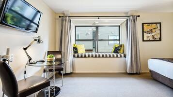 Una habitación de hotel con cama, escritorio con silla, televisor montado en la pared, ventana con cortinas y un cuadro en la pared.