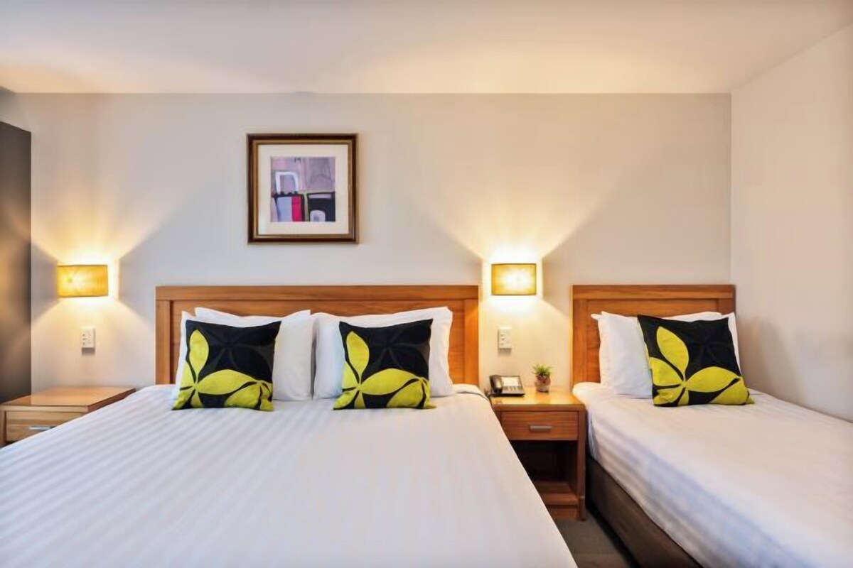 A hotel room with two beds, each with a wooden headboard, white bedding, and a single yellow and black pillow. There are bedside tables with lamps and a framed picture on the wall.