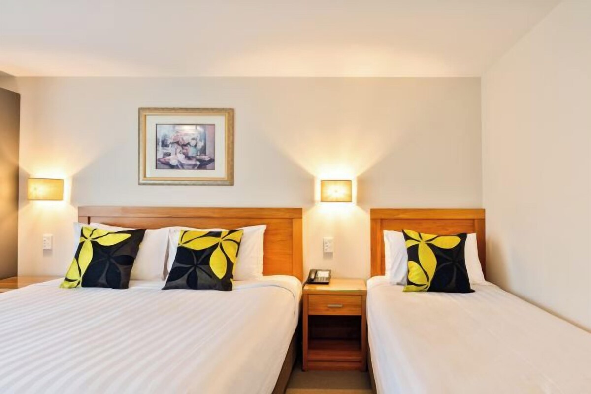 A hotel room with two beds, each with a wooden headboard, white linens, and decorative pillows. There is a framed picture on the wall and two bedside lamps.