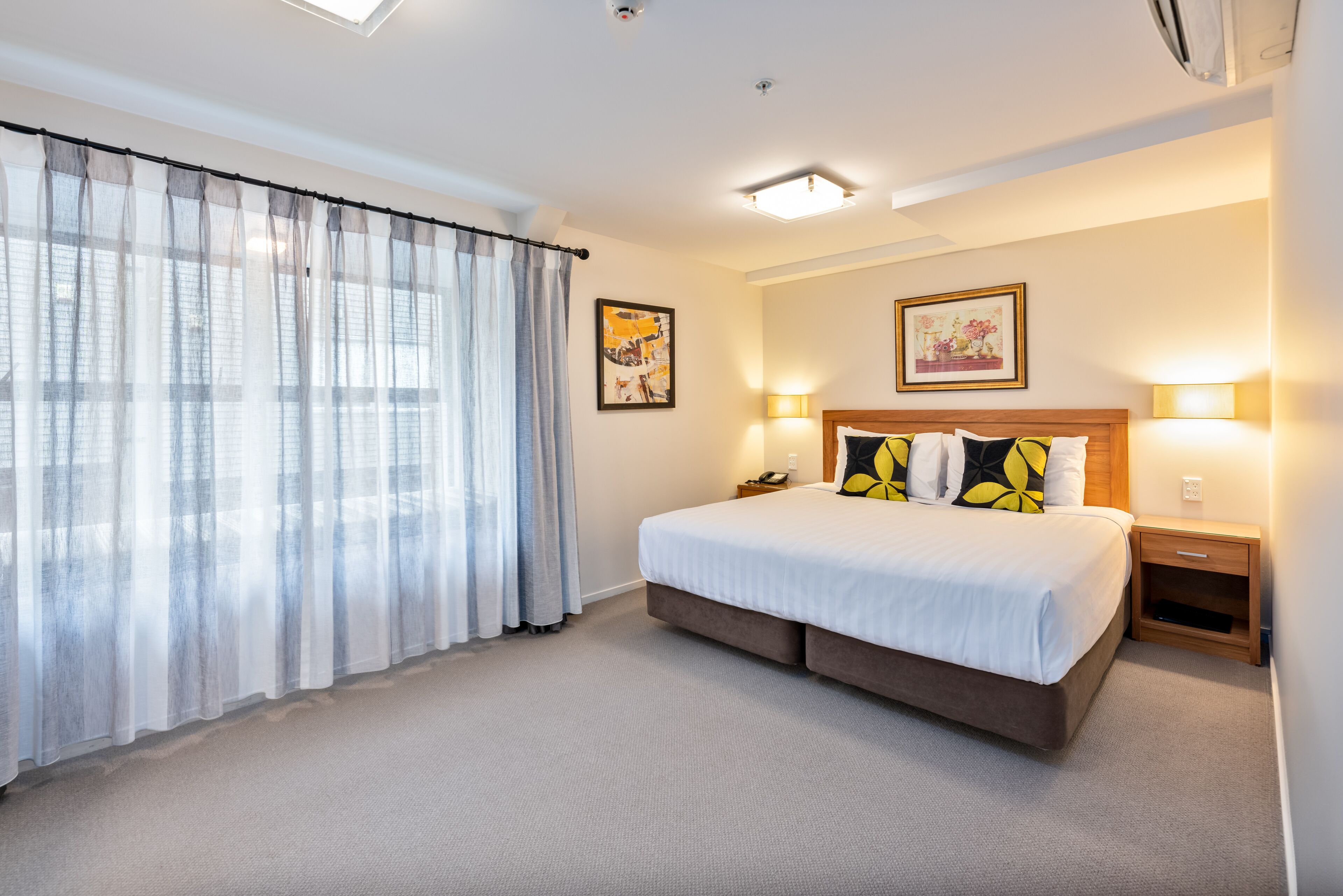 A hotel room with a large bed, a bedside table, a nightstand, a window with sheer curtains, and two wall-mounted lamps.