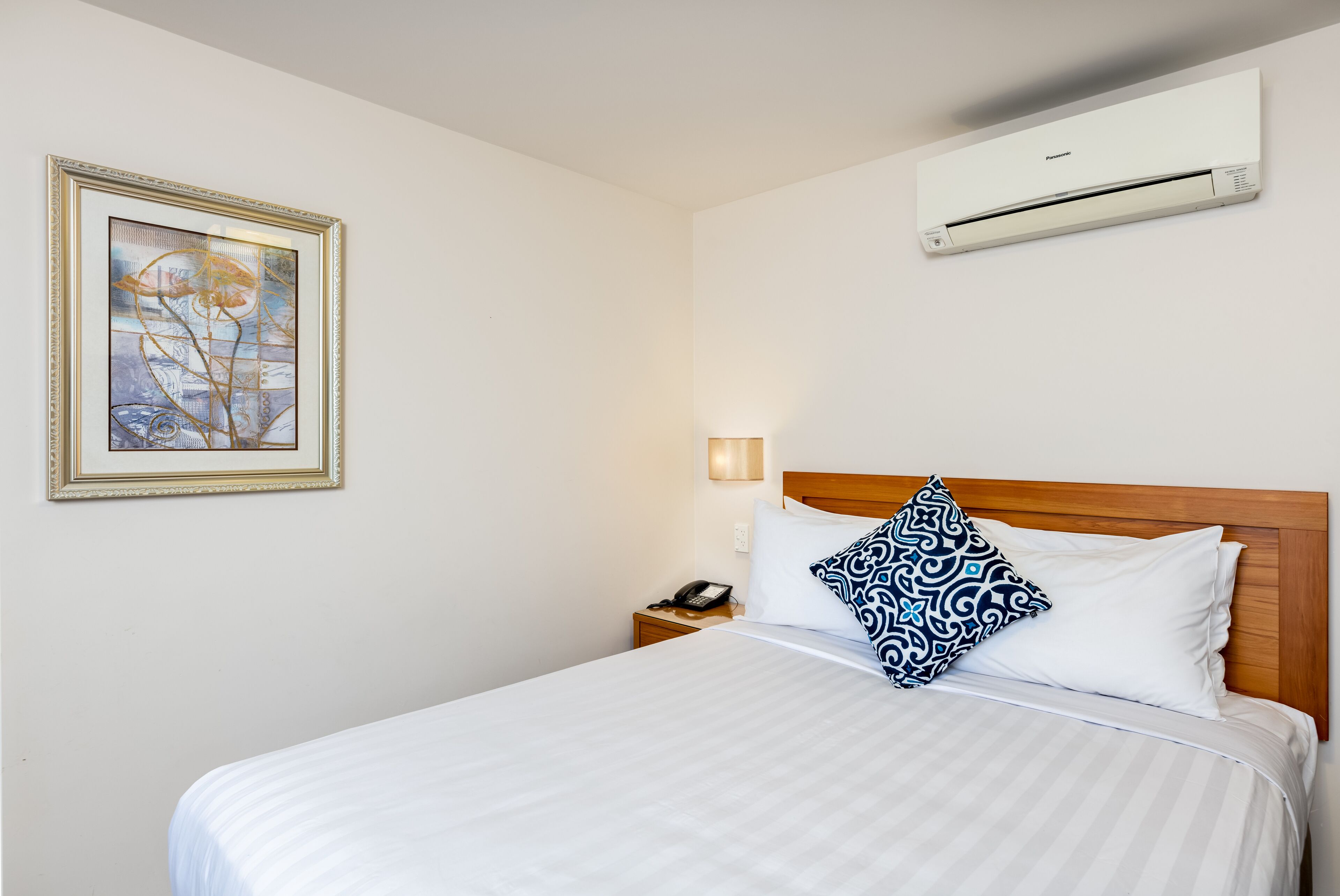 A hotel room with a bed, a nightstand with a phone, a wall-mounted air conditioner, and a framed artwork on the wall.