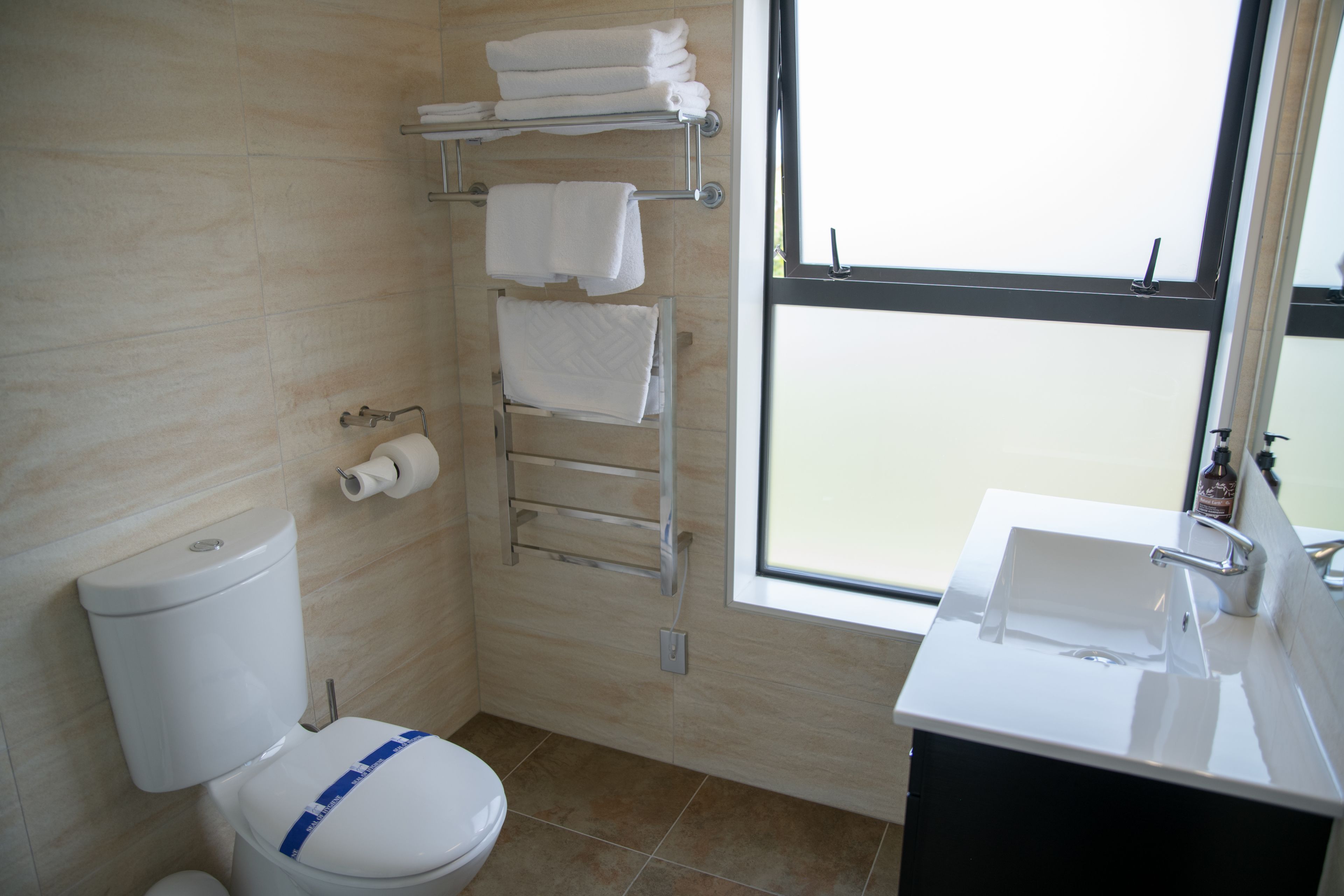 A bathroom with a toilet, a towel rack with towels, a sink, and a window.