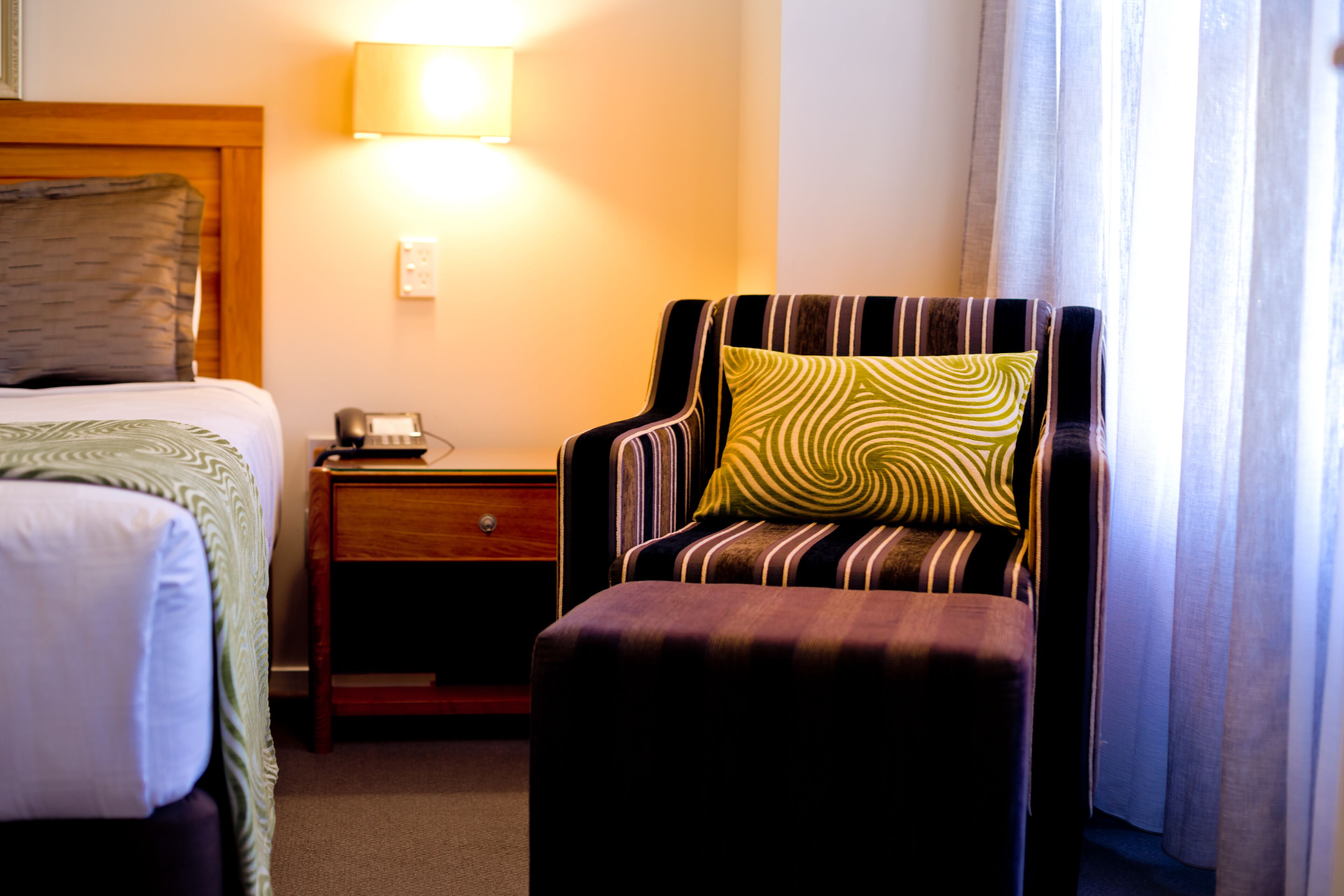 Une chambre d’hôtel avec un lit, une table de chevet, une chaise avec un coussin à rayures et une lampe.