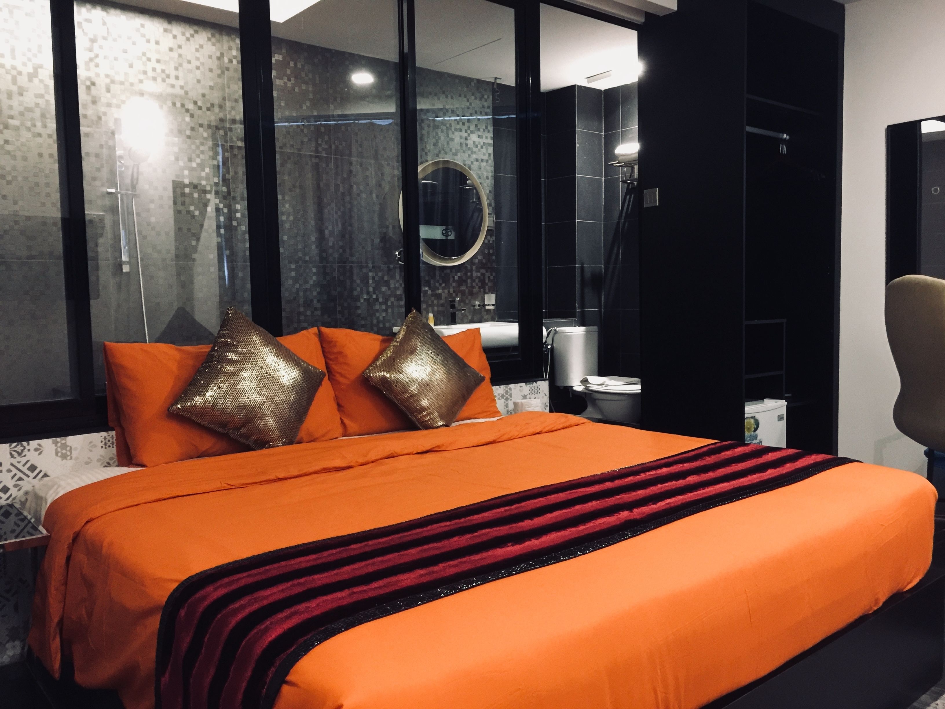 A modern hotel room with a large bed, orange bedding, a black and red striped throw, a large window, and a mirror above the bed.