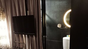 Habitación de hotel con cama, televisión, silla, baño con lavamanos y espejo, y vista al exterior.