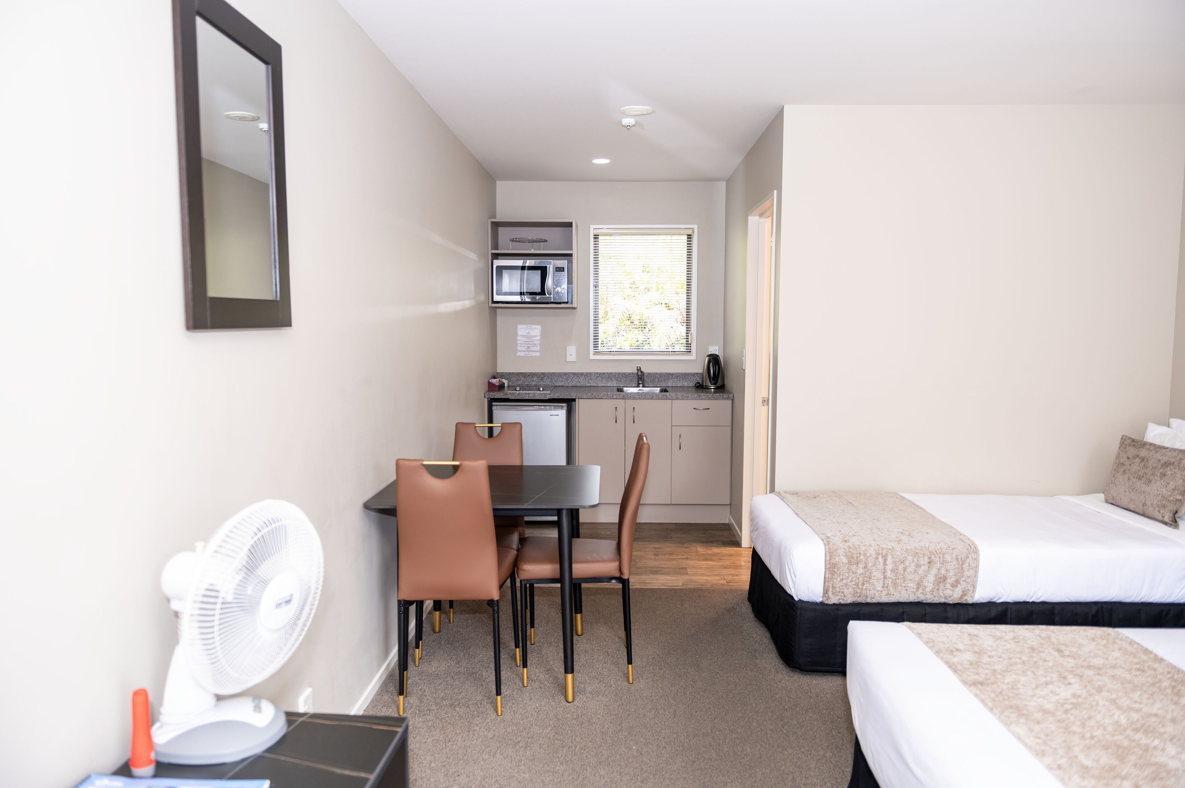 Een compacte hotelkamer met een bed, een klein bureau met stoel, een eettafel met stoelen, een kitchenette met oven en magnetron, en een raam met jaloezieën.