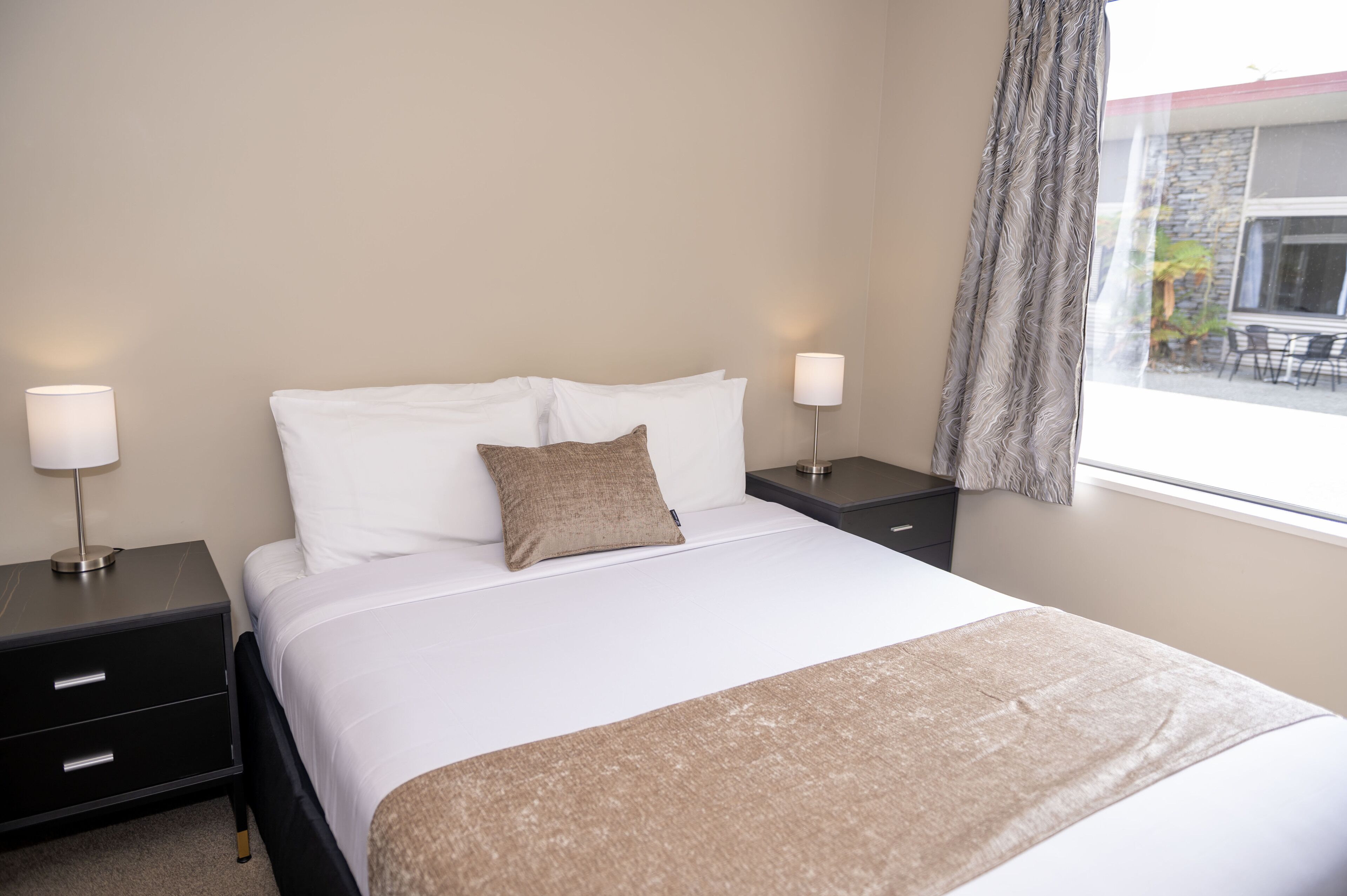 A neatly made bed with white linens and a beige throw blanket, flanked by two bedside tables with lamps, in a room with a large window and a view of the outside.