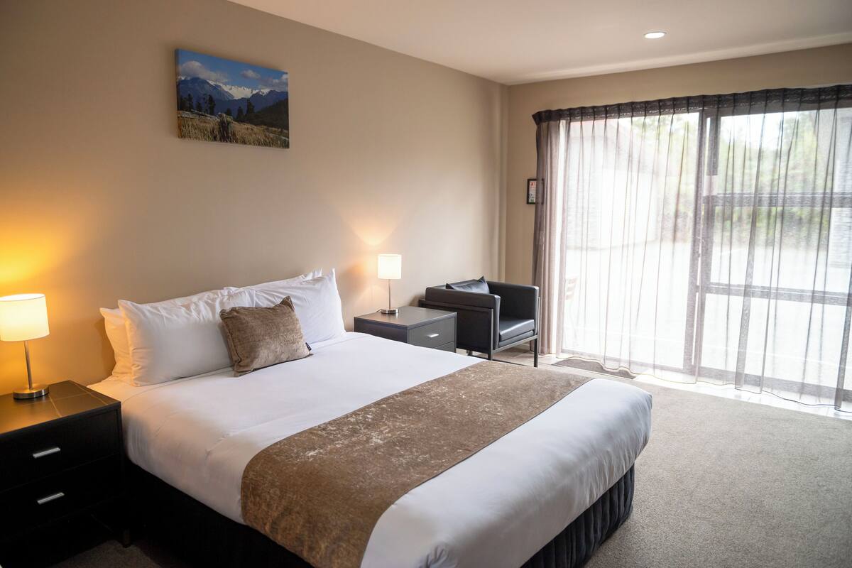 A hotel room with a large bed, a desk, a chair, a lamp, a picture on the wall, and a view through the window.