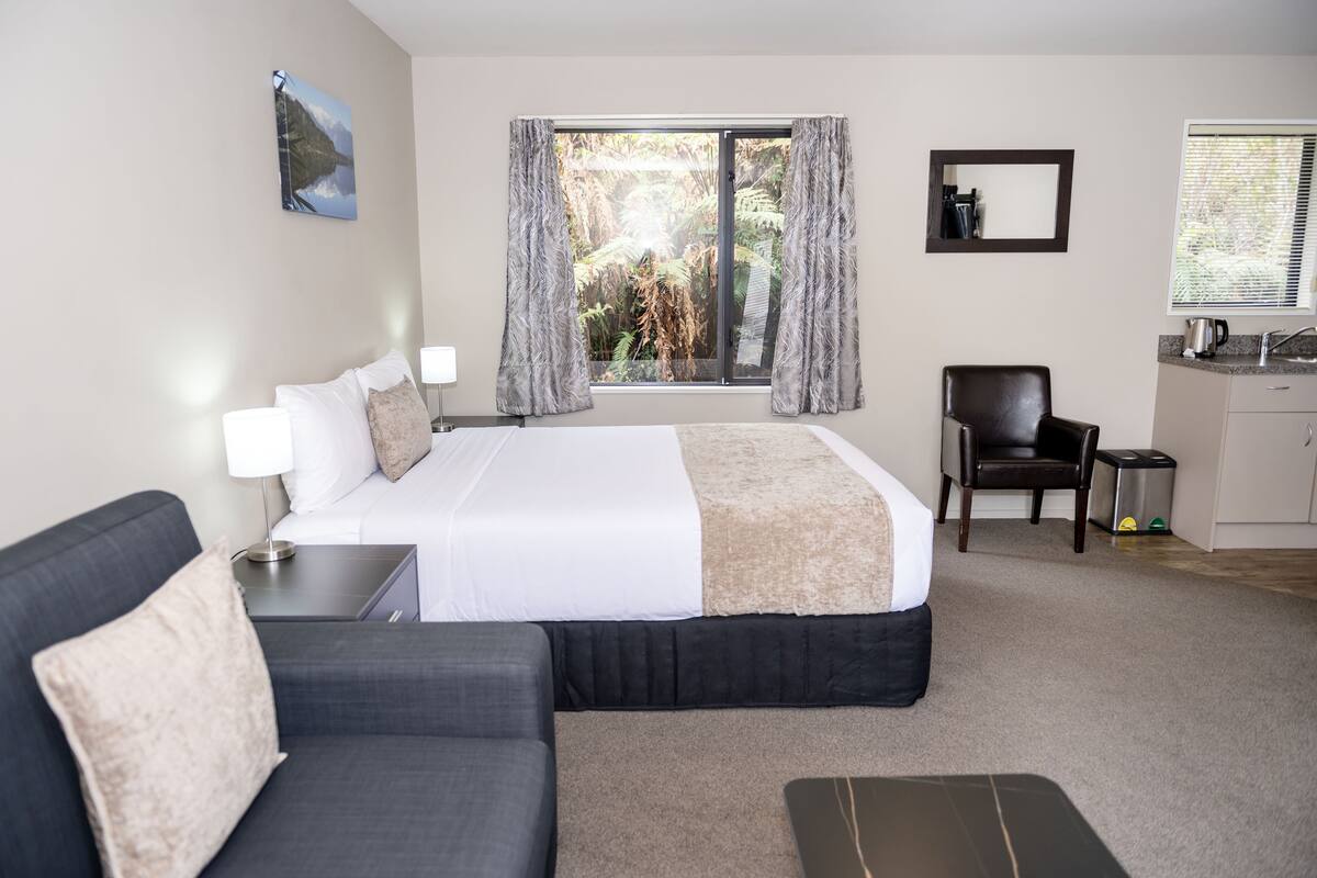 A hotel room with a bed, a desk, a chair, a small kitchenette, and a window with curtains.